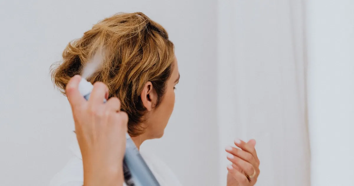 Une femme utilise de la laque pour coiffer ses cheveux dans un intérieur lumineux.