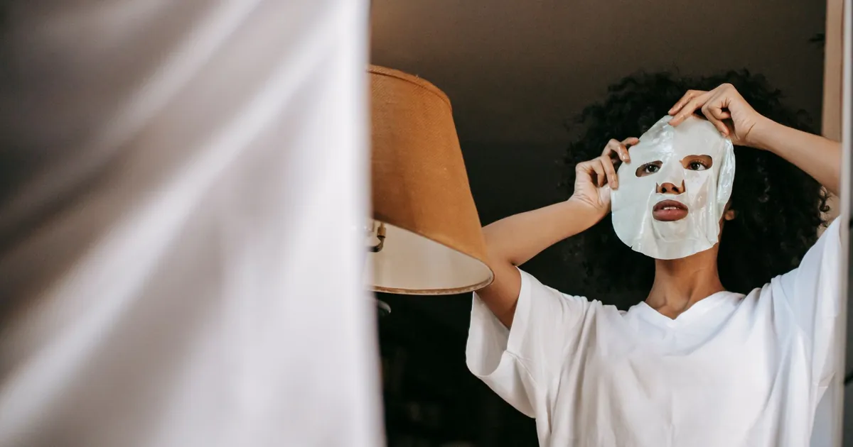 Vue en contre-plongée d'une femme noire aux cheveux bouclés appliquant un masque en tissu devant un miroir.