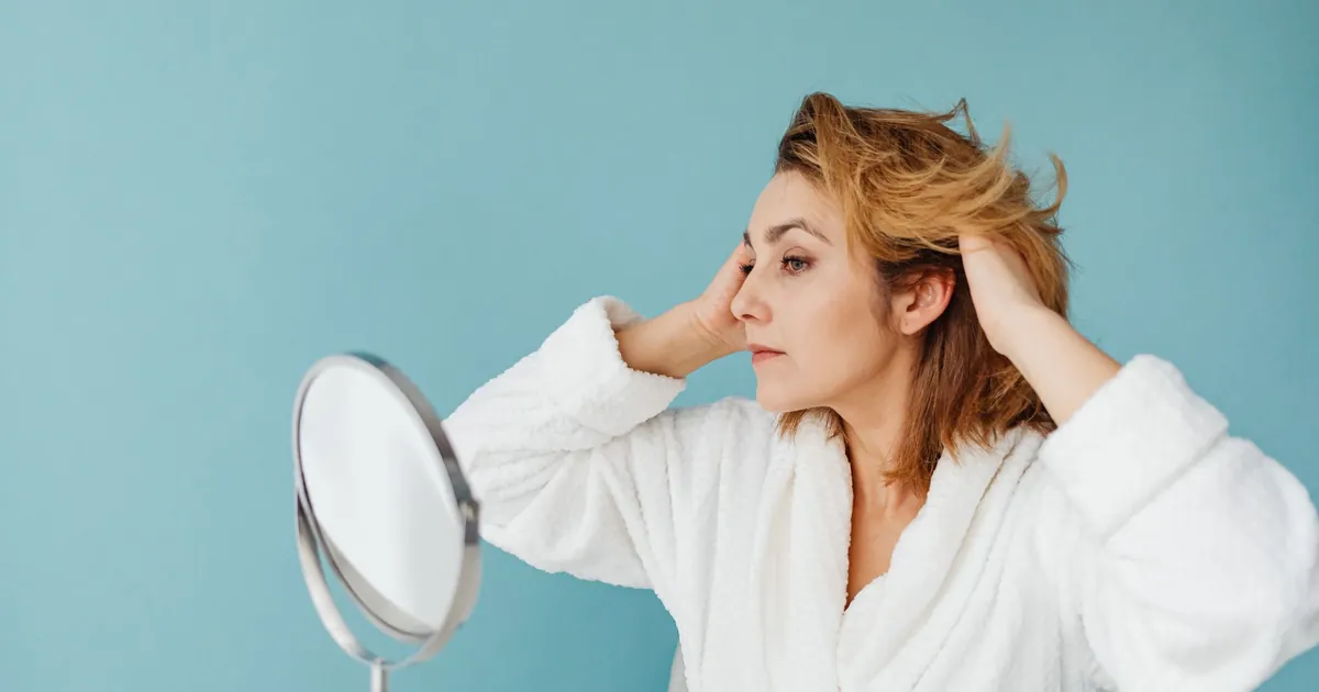Femme adulte en peignoir se coiffant devant un miroir sur fond bleu.