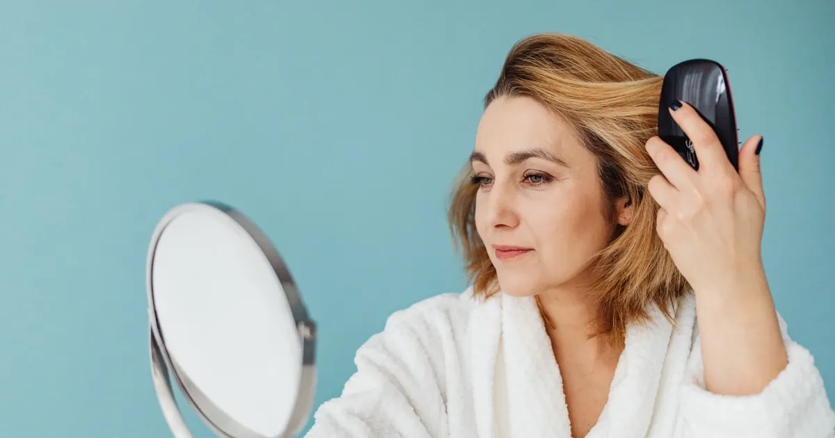Une femme en peignoir se peigne en regardant dans un miroir, expression calme.