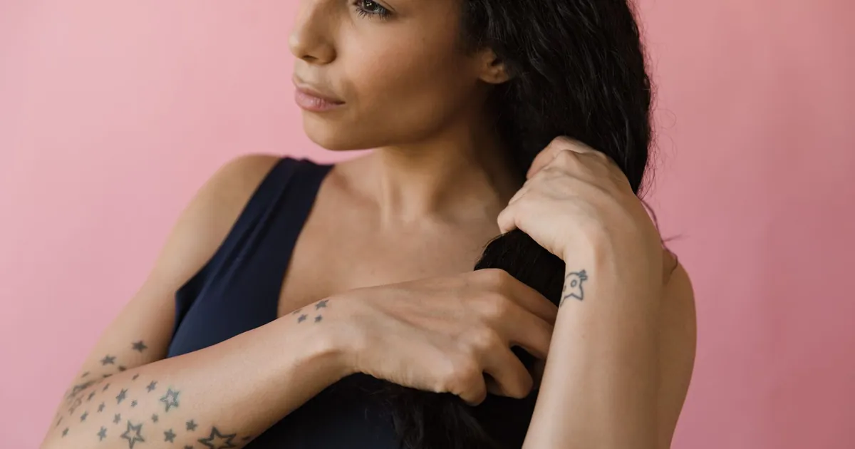 Femme tatouée ajustant ses cheveux sur fond rose.