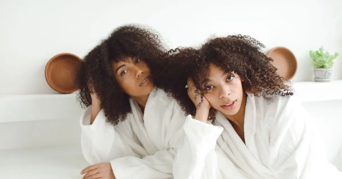 Deux femmes noires en peignoirs blancs aux cheveux bouclés, détendues à l'intérieur.