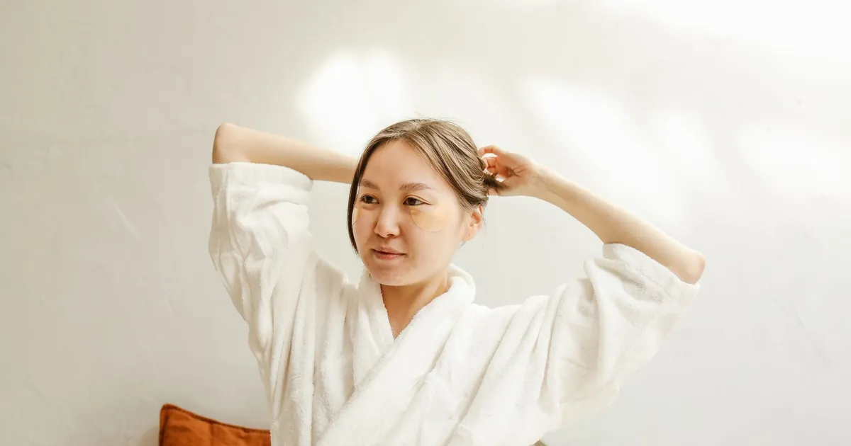 Femme asiatique en peignoir blanc appliquant des masques sous les yeux, intérieur lumineux.