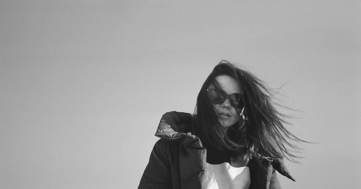 Portrait noir et blanc d'une femme dehors, cheveux au vent et lunettes de soleil.