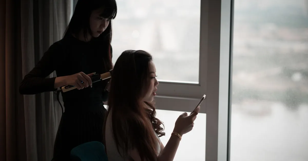 Femme asiatique bouclant ses cheveux en utilisant son smartphone près d'une fenêtre.