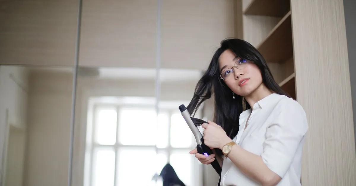 Femme asiatique bouclant ses cheveux au fer, regardant au loin le matin chez elle.
