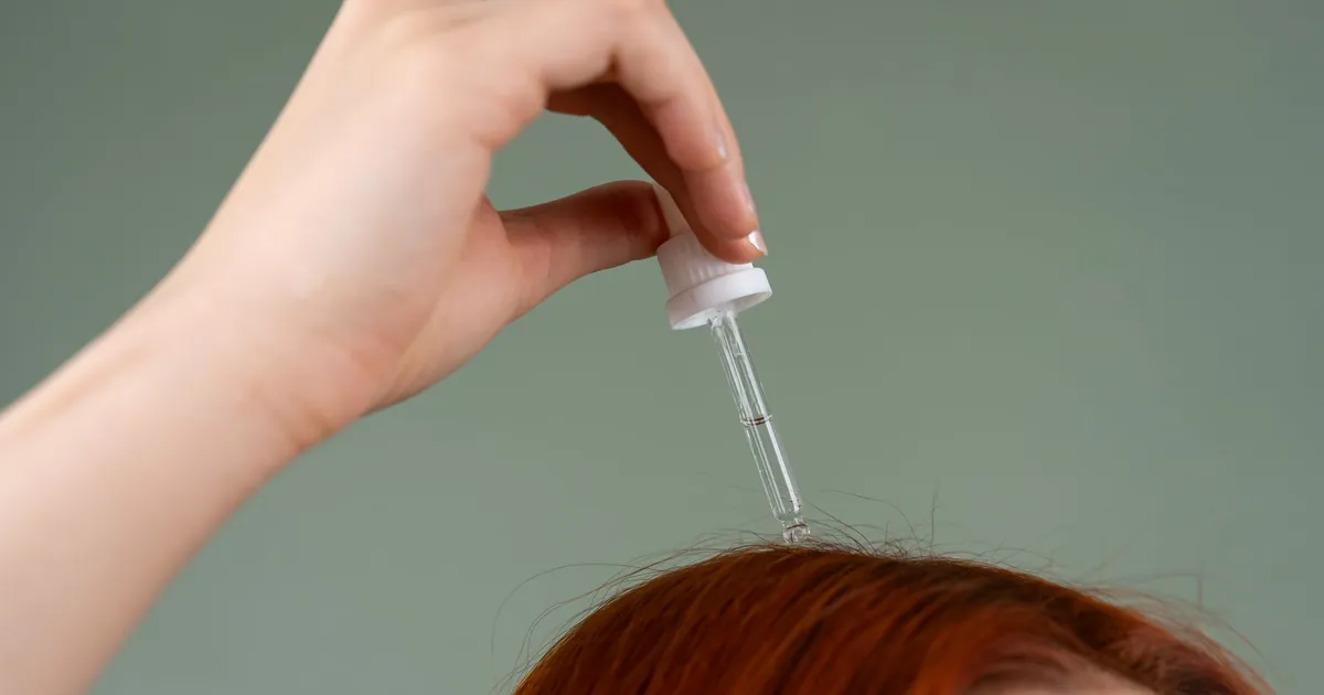 Main appliquant un sérum capillaire sur cheveux roux avec une pipette, gros plan.