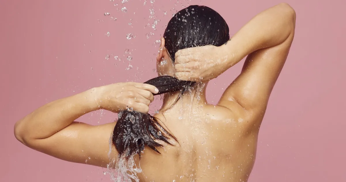 Femme aux longs cheveux se lavant sous la douche, gouttes d'eau sur fond rose.
