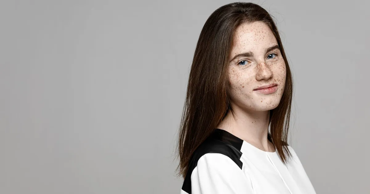 Jeune femme aux taches de rousseur et cheveux bruns, sourire calme sur fond gris